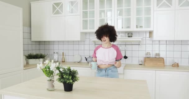 Heureuse jeune femme aux cheveux bouclés danse dans la cuisine en buvant un café . — Video
