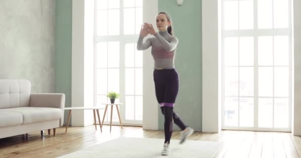 Deportiva mujer está entrenando haciendo sentadillas y embestidas en casa en la sala de estar . — Vídeos de Stock