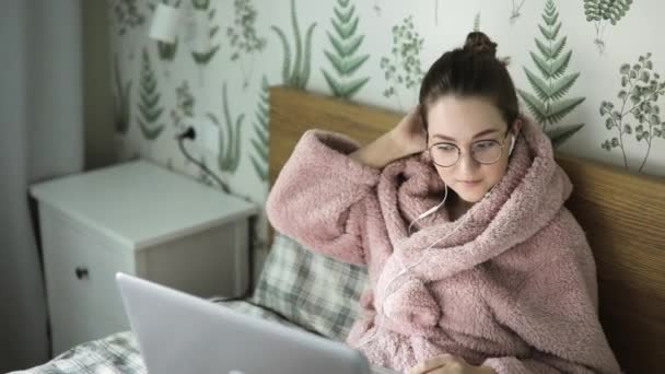 Muchacha joven en albornoz acostado en la cama usando el ordenador portátil para el trabajo . — Vídeos de Stock