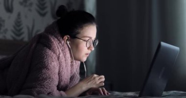 Kadın evde dizüstü bilgisayarla yatıyor. Kız öğrenci internet öğretmeniyle kulaklık çalışması yapıyor. Dil konuşmayı öğreniyor. Laptopa bakıyor.