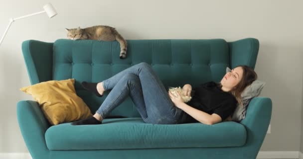 若い女性はソファに横になり、ガラスのボウルからポップコーンを食べます,自宅で休んで. — ストック動画