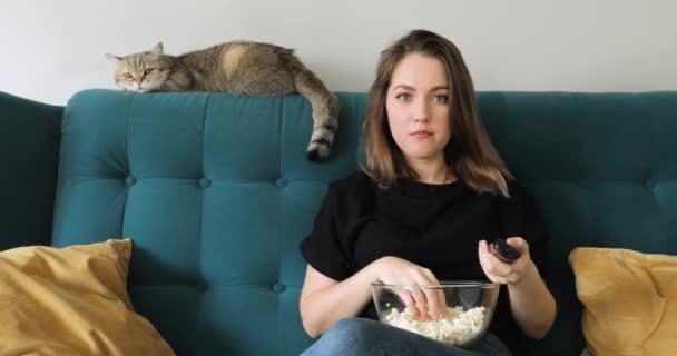 Vrouw eet popcorn zittend op de bank thuis schakelen kanalen afstandsbediening. — Stockvideo