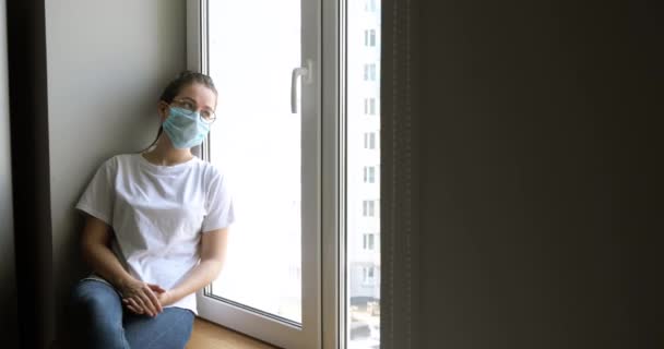 Vrouw met masker saai zittend op vensterbank en kijkend naar straat op quarantaine. — Stockvideo