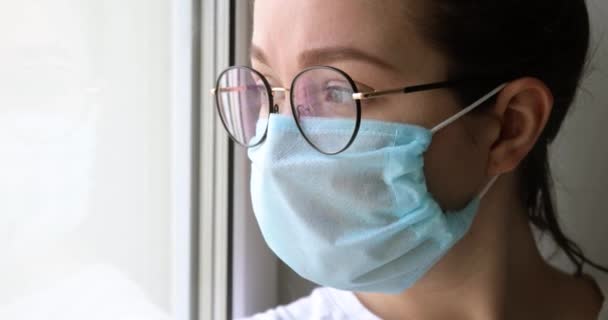 Melancholiek meisje in masker zit in de buurt venster saai op coronavirus quarantaine. — Stockvideo