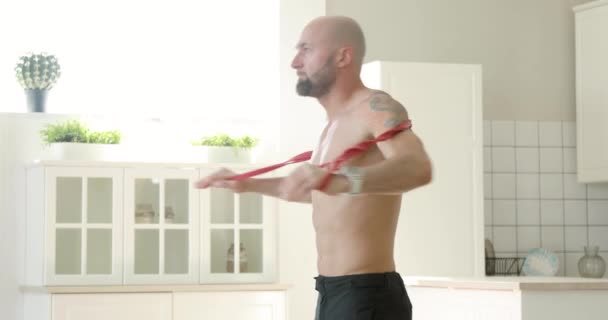 Man gör fitness motion för axlar med hjälp av elastiskt band hemma, sidovy. — Stockvideo