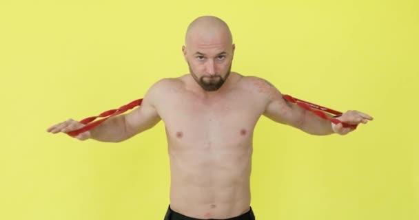 Man does fitness exercise for shoulders using elastic band on yellow background. — Stock Video