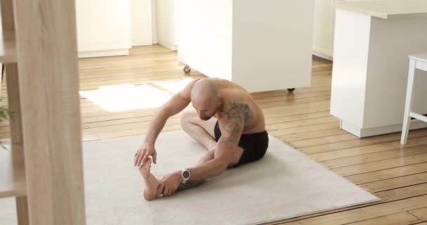 Muscular young man making stretching exercise sitting at home leans to legs. — Stock Video