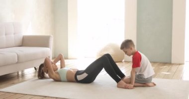 Kadın anne evde karın kasları yapıyor ve oğlu bacaklarını tutmasına yardım ediyor..