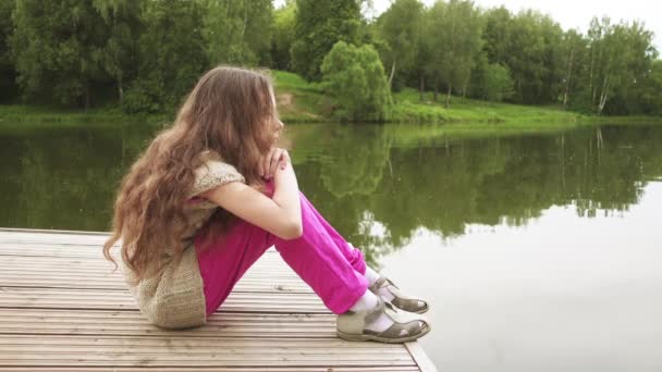 Mooi beetje verdrietig meisje zit op de houten brug in de buurt van het meer. — Stockvideo