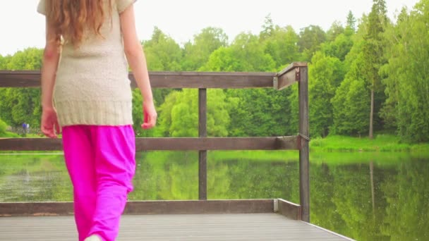 Hermosa niña caminando sobre un puente de madera sobre un fondo de lago de verano — Vídeos de Stock