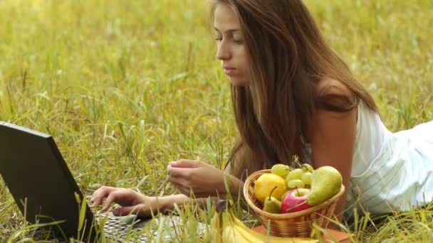 Красивая молодая женщина лежит на траве с ноутбуком и корзиной фруктов в летнем зеленом городском парке . — стоковое видео