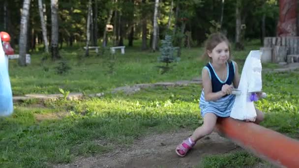 Piccola bella ragazza si siede su un'altalena di legno sullo sfondo di un parco cittadino estivo . — Video Stock