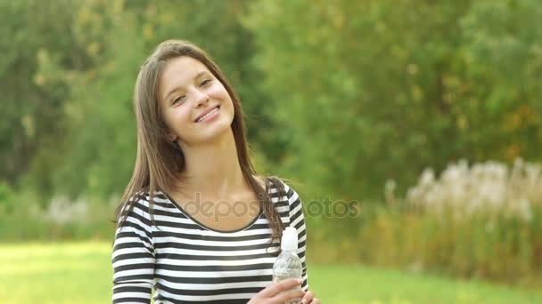 Красивая молодая женщина в полосатой куртке, стоящая в парке летом пьет воду из бутылки — стоковое видео