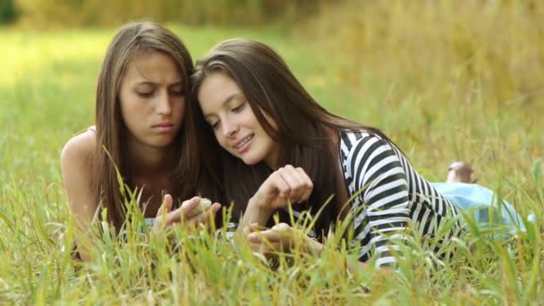 Dos hermosas mujeres jóvenes se encuentran en la hierba verde y leer sms en el teléfono móvil . — Vídeos de Stock