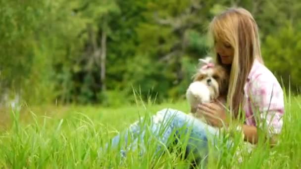 Νεαρή όμορφη γυναίκα κατέχει μικρό χνουδάτος σκύλοs στην αγκαλιά της και κάθεται στο γρασίδι. — Αρχείο Βίντεο