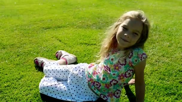 Liten vacker flicka sitter på grön gräsmatta i sommar stadspark. — Stockvideo