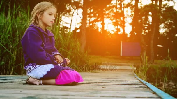 Small beautiful girl is sitting on wooden bridge near forest lake. — Stock Video