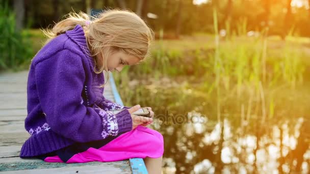 Jeune belle fille est assise sur un pont en bois près d'un lac forestier et lit un livre électronique . — Video