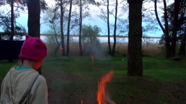 Small beautiful girl throws wood on fire in evening forest. — Stock Video