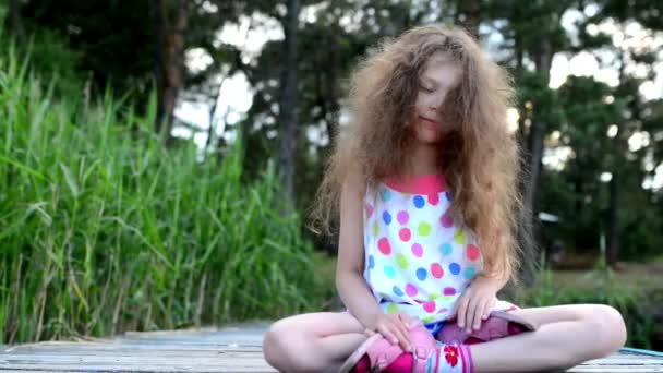 Pequeña chica hermosa en vestido multicolor está meditando en el puente de madera cerca del lago del bosque . — Vídeos de Stock
