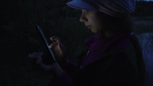 작은 아름 다운 여자는가 숲의 호수의 배경으로 황혼에 태블릿 컴퓨터에서 읽습니다.. — 비디오