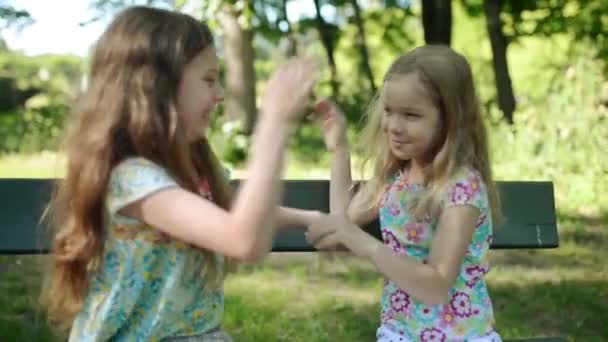 Two little beautiful sisters play on wooden bench in summer city park. — Stock Video