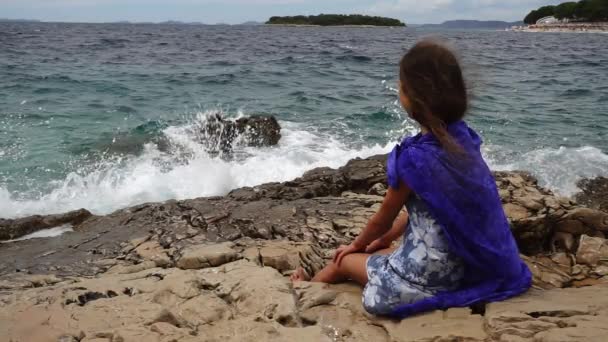 Malá krásná dívka sedí na skalách u mořského pobřeží a dívá do vzdálenosti. — Stock video