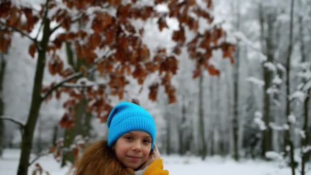 Pequena menina bonita em jaqueta quente senta-se no banco no parque de inverno coberto de neve . — Vídeo de Stock