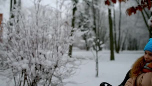 따뜻한 자 켓에 아름 다운 소녀 눈 덮인 겨울 공원에서 벤치에 앉아서 휴대 전화에서 재생. — 비디오