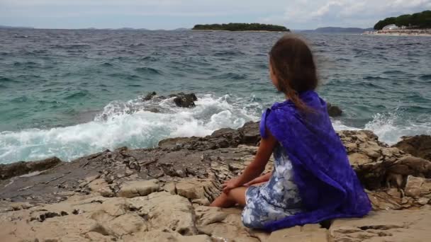 Lassú mozgás: gyönyörű kislány ül a sziklák közelében a tenger partján, és úgy néz ki, távolba. — Stock videók