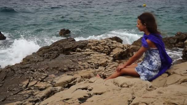 Movimento lento: a menina bonita pequena senta-se em rochas perto da costa do mar e olha para a distância . — Vídeo de Stock