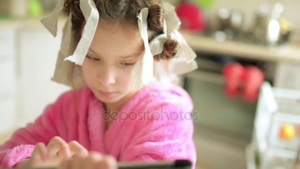 Kleine mooie meisje in roze badjas en rag krultang zit aan tafel in de keuken en speelt op tablet pc. — Stockvideo