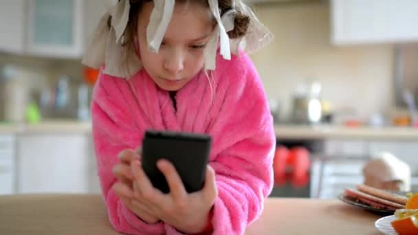 Mooie meisje in roze badjas en rag krultang zit aan tafel in de keuken en speelt op haar mobiele telefoon. — Stockvideo