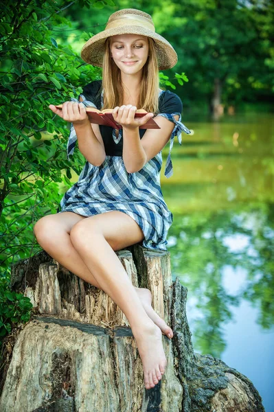 美しいです若いです若いです女性でわら帽子とともに本で彼女の手座っていますスタンプ近く夏の湖 — ストック写真