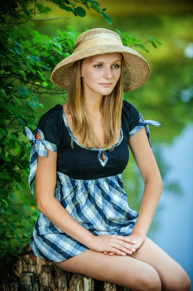 美しいです若いです女性でわら帽子に座っています夏の湖の近くの切り株 — ストック写真