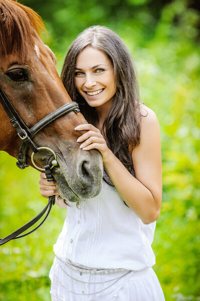 Woman in white dress keeps brown horse against background of summer green park.