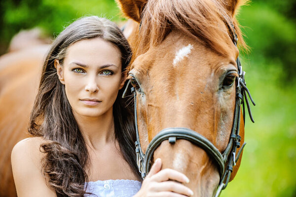 Woman in white dress keeps brown horse against background of summer green park.