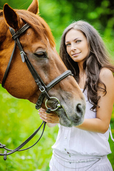 Woman in white dress keeps brown horse against background of summer green park.