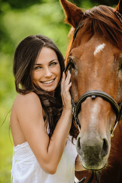 Woman in white dress keeps brown horse against background of summer green park.