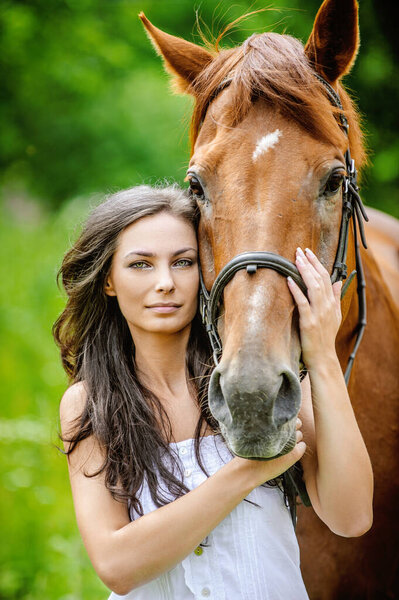 Woman in white dress keeps brown horse against background of summer green park.
