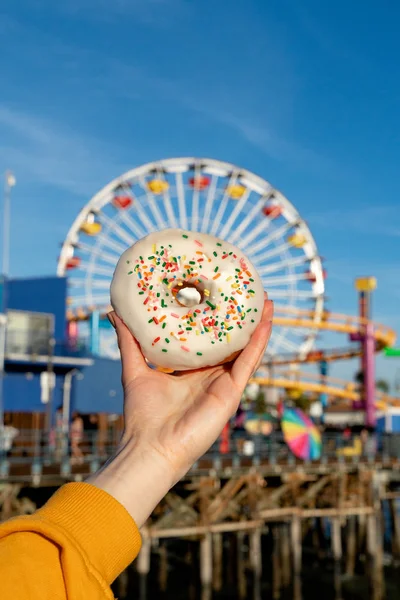 Female hand holding a donut on a background of a ferris wheel on — Zdjęcie stockowe