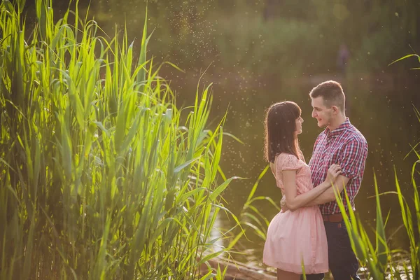 Milenci v jezeře. Mladý pár v lásce sedí v parku u vody, zatímco tito mladí muži hrají na kytaru v západu slunce. — Stock fotografie