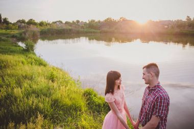 Göldeki aşıklar. Bu genç adam gün batımında gitar çalarken genç çift parkta, suyun kenarında oturuyor..