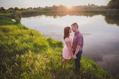 Göldeki aşıklar. Bu genç adam gün batımında gitar çalarken genç çift parkta, suyun kenarında oturuyor..
