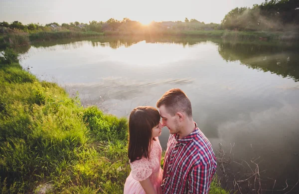 Amantes num lago. Jovem casal apaixonado sentado no chão do parque perto da água, enquanto estes jovens tocando guitarra ao pôr do sol. — Fotografia de Stock