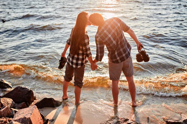 Любляча пара в картатих сорочках стоїть на березі озера, цілується і зустрічає захід сонця — стокове фото