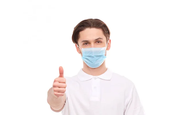 Ένας άντρας με λευκό μπλουζάκι ευρωπαϊκής εμφάνισης με ιατρική μάσκα, δείχνει τον αντίχειρά του. Στη μάσκα είστε προστατευµένοι από ιούς covid 19. — Φωτογραφία Αρχείου