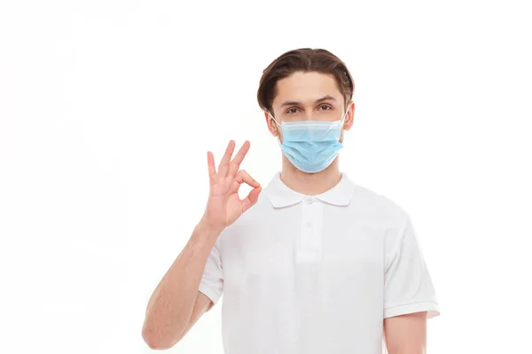Un hombre con una camiseta blanca de apariencia europea con una máscara médica, muestra un pulgar hacia arriba. En la máscara está protegido de virus covid 19 . — Foto de Stock