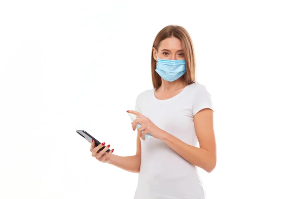 Mujer joven con máscara médica lava su teléfono móvil con un aerosol antiséptico de gel. Foto conceptual sobre el tema de la epidemia . — Foto de Stock