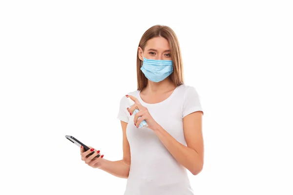 Mujer joven con máscara médica lava su teléfono móvil con un aerosol antiséptico de gel. Foto conceptual sobre el tema de la epidemia . — Foto de Stock